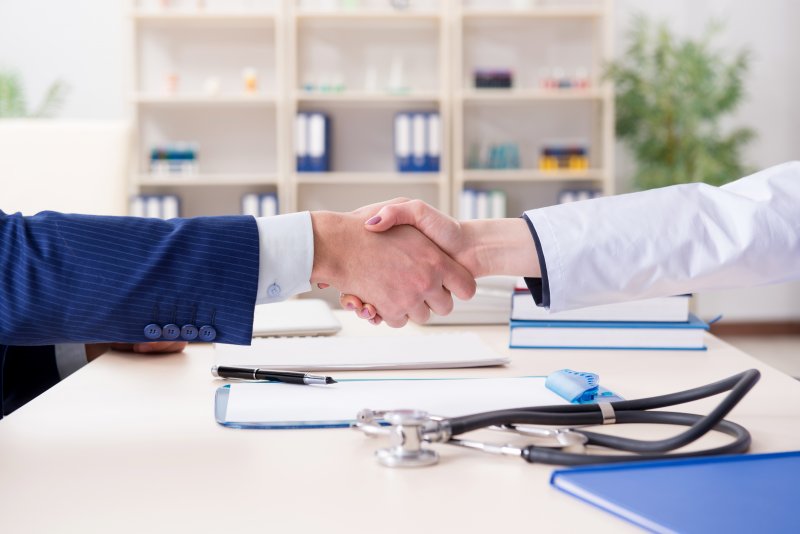 Two people shaking hands across a table that contains paperwork, a stethoscope, and books